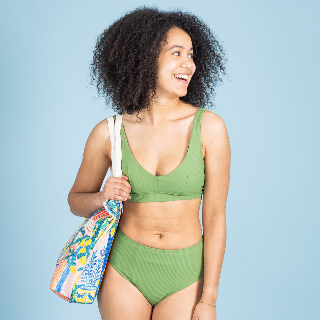 Model draagt de groene STELLA menstruatiebikinislip van Moi et Marie met bijpassende zwemtop en kleurrijke strandtas.